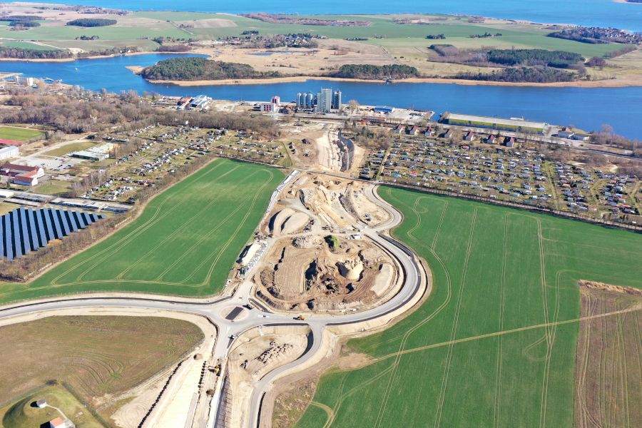 Auf einem Feld befindet sich eine riesige Baustelle, auf der Bagger, Radlader und Lkw Erde bewegen. Im Hintergrund sind die Stadt Wolgast, der Fluss Peene und Fabrikgebäude.