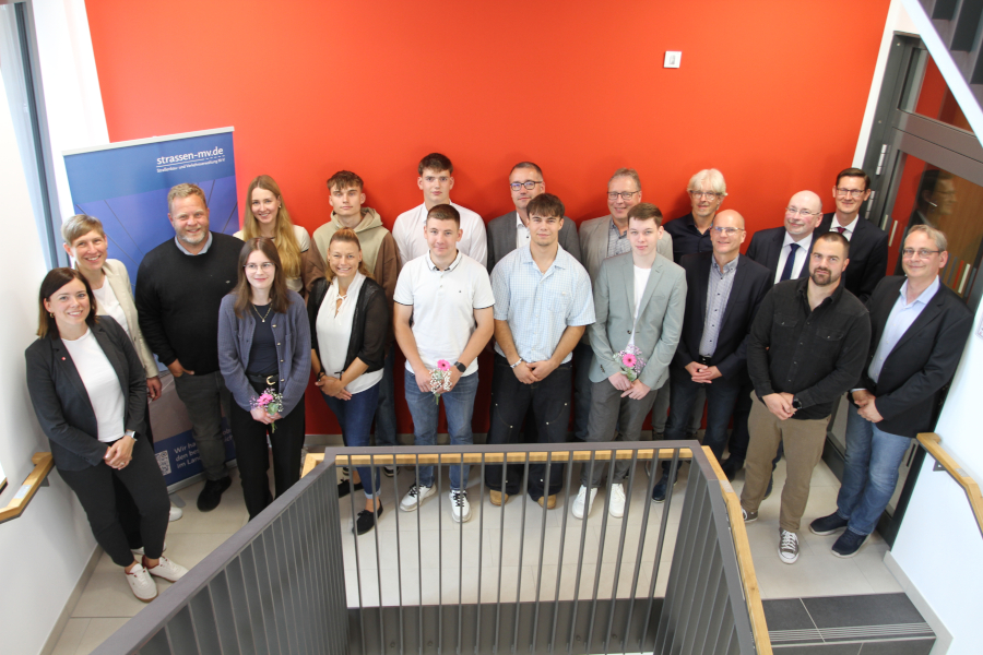 Gruppenfoto vor roter Wand: 20 Personen – darunter Auszubildende, Ausbilderinnen und Vertreterinnen des Straßenbauamts – stehen auf einem Treppenabsatz und lächeln in die Kamera. Einige halten Blumensträuße in der Hand. Im Hintergrund ein Roll-Up mit dem Logo 'strassen-mv.de'. © strassen-mv.de Gruppenfoto vor roter Wand: 20 Personen – darunter Auszubildende, Ausbilderinnen und Vertreterinnen des Straßenbauamts – stehen auf einem Treppenabsatz und lächeln in die Kamera. Einige halten Blumensträuße in der Hand. Im Hintergrund ein Roll-Up mit dem Logo 'strassen-mv.de'.